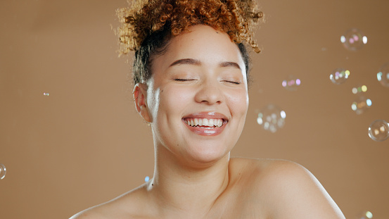 Woman, bubbles and skincare for beauty, happy with natural cosmetics and glow isolated on brown background. Dermatology, wellness for skin and liquid soap with self care and anti aging in studio