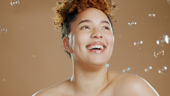 Happy woman, bubbles and skin for beauty, happy with natural cosmetics and glow on brown background. Dermatology, wellness with skincare and liquid soap for self care and anti aging in studio
