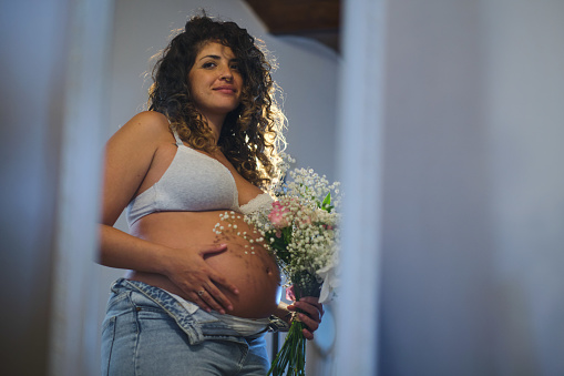 side view cozy portrait of a carefree latino woman with a positive attitude touching her belly indoors with a bouquet of flowers on her hand
