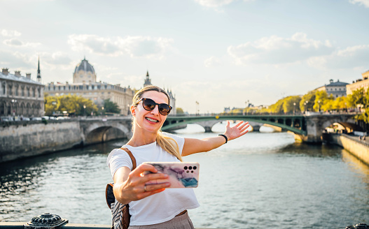Young happy woman standing on the bridge over the River Seine and making a vlog in Paris, France
