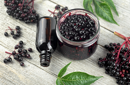Homemade black elderberry syrup in glass jar and bunches of black elderberry with green leaves on wooden desk.
