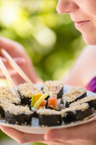 Young woman, lower part of the face, eating sushi with shopsticks from plate, full of  maki sushi and smilling. Bright green background.