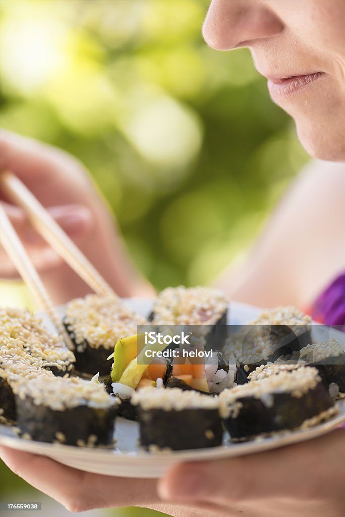 young woman eating sushi Young woman, lower part of the face, eating sushi with shopsticks from plate, full of  maki sushi and smilling. Bright green background. 20-24 Years Stock Photo