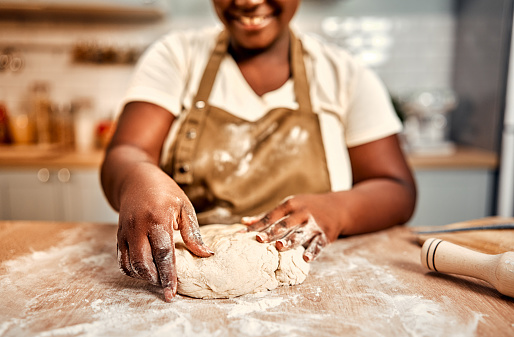 Close up of smiling curvy woman in brown apron shaping raw dough in flour on wooden kitchen table. African american lady preparing traditional homemade bread during daily routine.