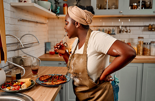 Plus size african american woman trying with fork tasty traditional dish prepared on modern home kitchen. Side view of hostess in turban and apron making first taste before serving.