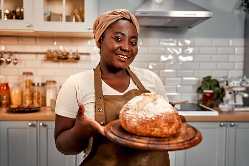 Happy hostess in headband and apron holding freshly baked crunchy bread on wooden board. Smiling curvy woman with black skin proudly looking at camera while standing on home kitchen.