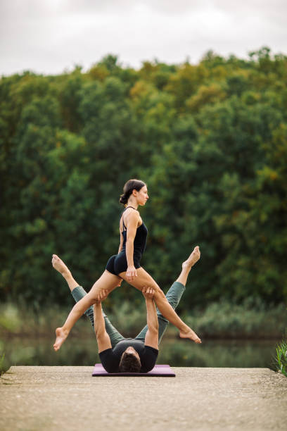 Young man and woman doing acroyoga by lake Young man and woman doing acroyoga pose by lake, strength, trust, perfect synchronization, balance and harmony, forest in the background acroyoga stock pictures, royalty-free photos & images