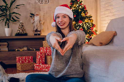 A beautiful woman sits in front of a Christmas tree and shows her heart with her hands