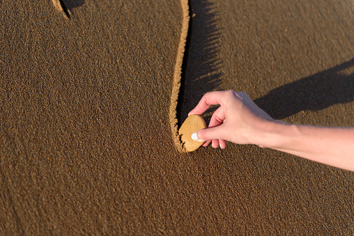 Young woman on the beach is drawing a heart shape on the sand at sunset.