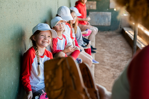 Multi-ethnic girls (9 years) on softball team at the ball park, sitting in dugout.