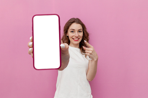 young cute woman in white t-shirt shows blank smartphone screen on pink isolated background, the girl advertises copy space on the phone, mock-up