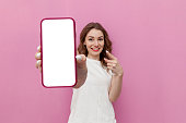 young cute woman in white t-shirt shows blank smartphone screen on pink isolated background