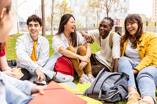 Young group of multiracial students laughing and having fun together sitting on the grass in the university campus. Teenage classmates social gathering relaxing outdoors.