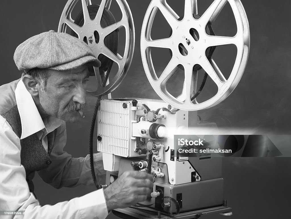 Senior Man Projectionist Starting Film With Old Fashioned Film Projector Senior man wearing shirt,waist and flat cap starting old fashioned film projector.He is placed on the left side of the frame.Smoke ad light bean come out of projector lens.The photo is black and white and has horizontal composition.Shot with a Full frame DSLR camera.  Movie Theater Stock Photo