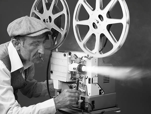 Senior Man Projectionist Starting Film With Old Fashioned Film Projector Senior man wearing shirt,waist and flat cap starting old fashioned film projector.He is placed on the left side of the frame.Smoke ad light bean come out of projector lens.The photo is black and white and has horizontal composition.Shot with a Full frame DSLR camera.  television camera photos stock pictures, royalty-free photos & images