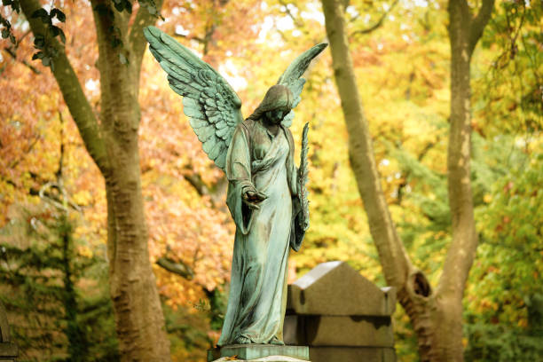an antique angel figure with a palm branch stands on a grave of a cemetery in front of an autumnal colorful leafy forest in a blurred background