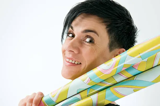Head-and-shoulder view of a black-haired middle-aged woman keeps smiling yellow/green patterned gift paper rolls over the shoulder friendly in the camera