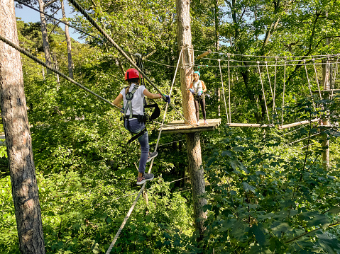 Happy school girl enjoying activity in a climbing adventure park on a summer day.Summer fun and sports for adventurous people.Teenager Girl walks a rope bridge between trees in an amusement park in safety gear and a helmet. Rope park in wood forest.