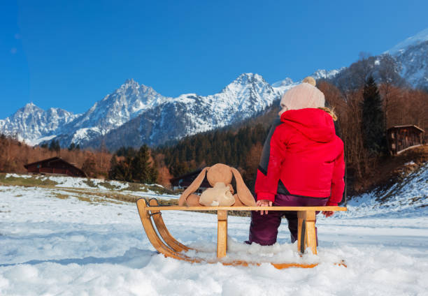 Little girl with teddy rabbit in the winter outfit sit on wooden sledge over high mountain range view from behind