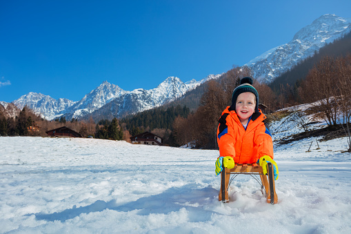 Smiling little child boy in winter orange coat and woolen hat lay on the sledge sliding downhill on the snow