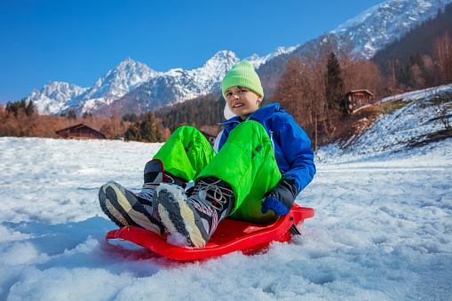 Young boy in winter outfit slide downhill on alpine slope in the mountains on red sledge