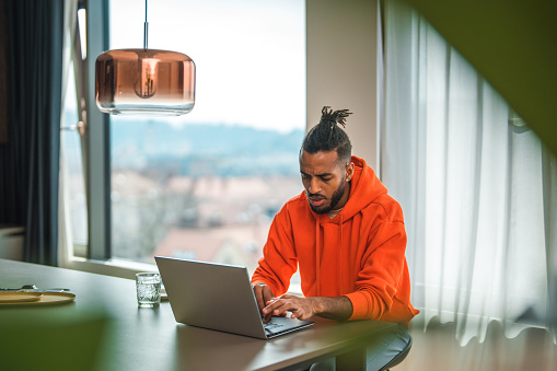 Front shot of black man using a laptop for work. He is seated at a modern home while wearing an orange sweater.