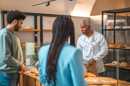 Diverse people standing in line in a small artisan bakery. Buying freshly baked goods. Mature Hispanic male shop assistant handing him his order.