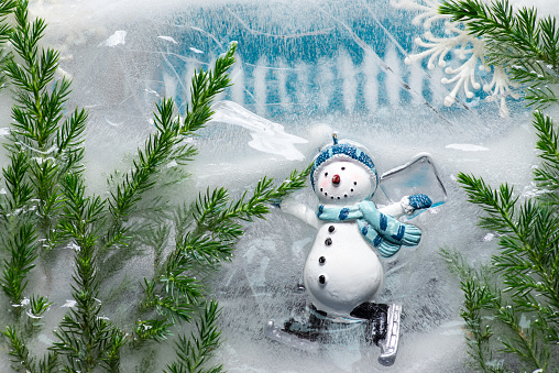 Winter, New Year or Christmas background with a Snowman dressed in a blue hat, scarf and gloves, with ice skates. The snowman is in a winter landscape, with snow, sparkling ice and green needles. Winter concept, Christmas and New Year atmosphere