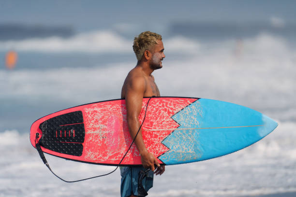 indonesischer männlicher surfer geht nach dem surfen mit surfbrett aus dem wasser - bali male beautiful ethnicity stock-fotos und bilder