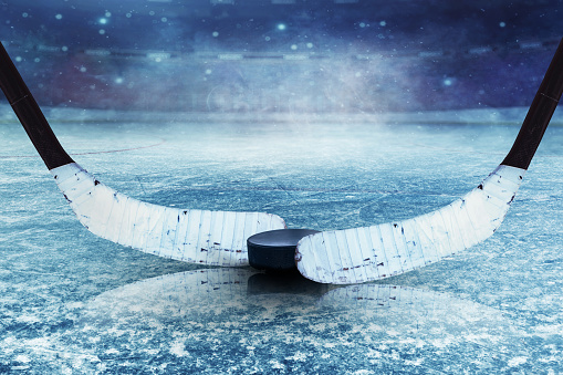 This high-quality stock photo shows two ice hockey sticks and a puck resting on the ice, with a hockey stadium in the background. The sticks are crossed together, and the puck is lying between them. The ice is smooth and clear, and the stadium is brightly lit.