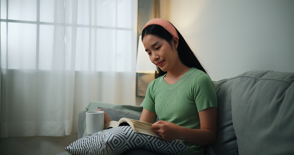 Portrait of Happy young Asian woman reading a book while sitting on sofa in the living room,relaxation lifestyle of single life people