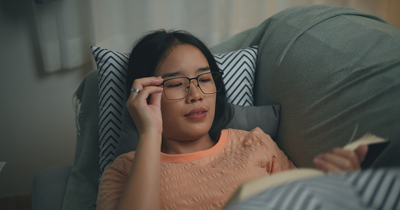 Portrait of Happy young Asian woman wearing glasses lying on sofa enjoy reading a book in the living room,relaxation lifestyle of single life people