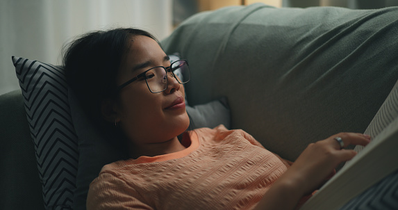 istock Young single woman wearing glasses reading a book on comfortable sofa. 1766507329