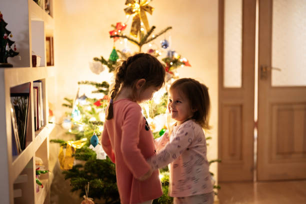 Two sisters together near Christmas tree at evening home.