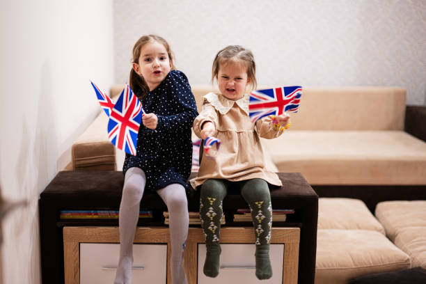 istock Two sisters are sitting on a couch at home with british flags on hands. Great Britain children girls with flag . 1766542856
