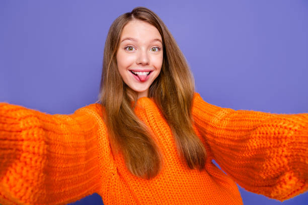 foto de cabelo castanho adolescente blogando menina vestindo elegante suéter quente laranja ela faz selfie mostrar língua isolada no fundo da cor violeta - smiling humor child making a face - fotografias e filmes do acervo