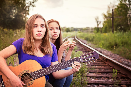 Two teenagers are sitting on railroad tracks during a vigil. One is strumming a guitar and softly singing while the other is holding a candle and reminiscing about the past.