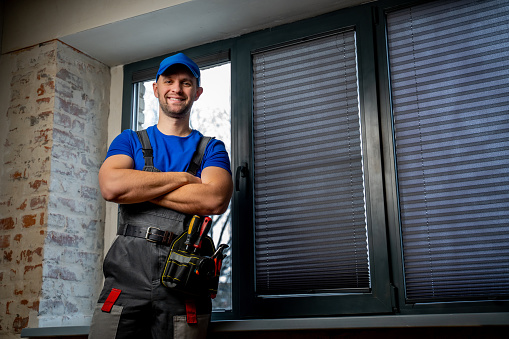 Maintenance worker in uniform, blue T-shirt and cap, crossing his arms. A handyman wearing a tool belt against a black window background.