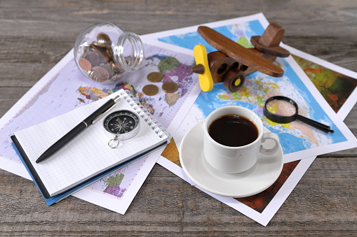 A white cup of coffee on the wooden background is next to world maps, compass, magnifier,  notebook, retro model of airplane and glass jar of coins. Concepts of  travel, tourism, choice and planning.