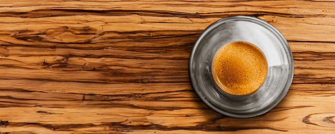 Top view of a glass cup of freshly brewed black espresso coffee with crema and golden fine bubble foam on a wood grain table. Barista art concept. Web banner.