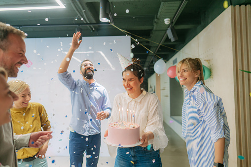 Photo of a group of coworkers celebrating birthday of their colleague - tossing confetti and balloons in the air, blowing candles on the cake and dancing