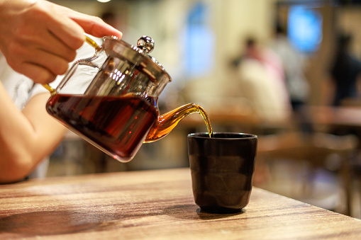 Hand pouring a cup of tea from a stylish transparent teapot into a tea cup in a restaurant