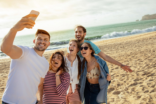 Happy millennial european and arabic friends have fun, taking selfie on smartphone, enjoy free time on ocean beach. App for blog and social network, outdoor party, holiday and trip