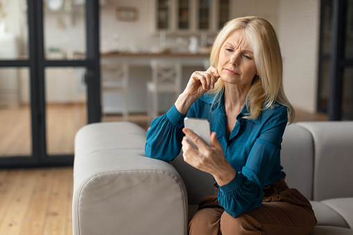 Bad news concept. Confused mature woman holding smartphone, looking at cellphone screen with worried expression, reading unplesant message, sitting on couch, copy space