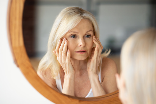 Aged skin problems. Upset senior woman looking at mirror and touching face, lady noticed wrinkles near eyes, unhappy with bad skincare routine and treatment