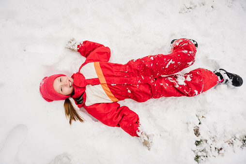 Girl falls into snow in mountain country in snowy forest. Little child in winter clothes having fun. Kid wearing warm hat lying down in winter park in holidays. Baby play ingamong snowdrifts. Top view