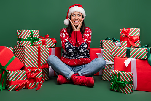 Full length cadre of astonished young lady hands cheeks wear red ornament sweater much christmas boxes isolated on green color background.