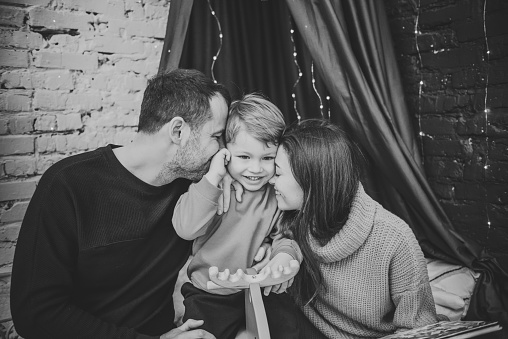 Concept of family holiday. Mother, father hug son at home. Happy New Year and Merry Christmas. Mom, dad embrace the kid. Christmas tree, decorated interior. Black and white photo closeup.