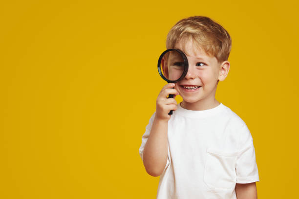 Cheerful little boy with blonde hair smiling while looking at free empty space for text through magnifier, standing against yellow background.