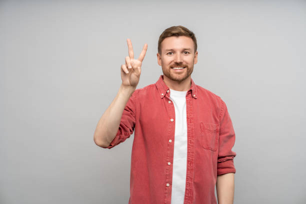 istock Smiling positive man showing victory sign looking at camera on gray background. 1766784495